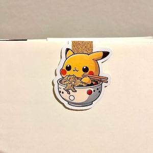 May include: A yellow Pikachu sticker eating ramen from a white bowl with red accents. The sticker features a cartoon Pikachu with red cheeks, black eyes, and a smiling expression. Chopsticks are visible. The ramen bowl is filled with noodles and vegetables.