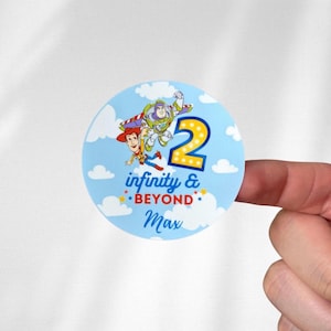 May include: A round sticker with a light blue background featuring cartoon characters Woody and Buzz Lightyear from Toy Story. The number 2 is in yellow, with the text "infinity & BEYOND" and a name below.