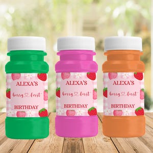 May include: Three colorful bubble bottles: green, pink, and orange. Each bottle has a white cap and a label with the text "ALEXA'S berry first BIRTHDAY" with strawberry and flower graphics. The bottles are on a wooden surface.