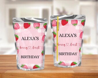 May include: Two silver pouches with a pink gingham pattern and strawberry illustrations. Text reads "ALEXA'S berry first BIRTHDAY". One pouch has a yellow straw. Suitable for a first birthday celebration.