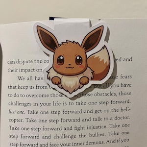 May include: A bookmark featuring a cartoon Eevee character. The bookmark is a sticker with a white border, showcasing Eevee's brown fur, ears, and tail. The bookmark is placed on a book with printed text.