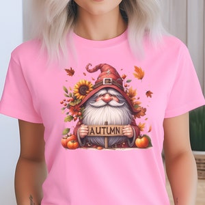 May include: A pink t-shirt with a graphic of a gnome holding a sign that says "AUTUMN". The gnome is wearing a red hat and has a white beard. The graphic is surrounded by fall leaves, sunflowers, and pumpkins.