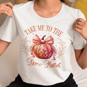 以下が含まれることがあります： 「Take Me To The Disco Patch」と書かれたグラフィックの白いTシャツ。グラフィックは、ピンクのリボンと葉が付いたディスコボールが特徴です。シャツの下部には「Tee Tinker」と「White」の文字も表示されています。