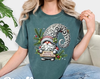 May include: Blue Spruce t-shirt featuring a Christmas gnome design. The gnome has a long white beard, a red and white hat, and holds a sign that reads "MERRY CHRISTMAS". The design includes decorative swirls and greenery.