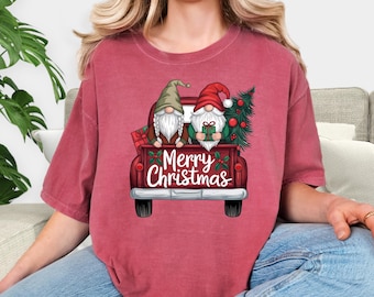May include: Crimson-colored t-shirt featuring a graphic of two gnomes in a red truck bed with a Christmas tree and gifts. The text "Merry Christmas" is displayed below the gnomes. The shirt is a comfortable, casual style.