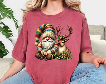 May include: Crimson-colored t-shirt featuring a whimsical Christmas gnome and a reindeer with holly antlers. The gnome wears a colorful striped hat and sweater. The reindeer is adorned with holly and sits among festive decorations.