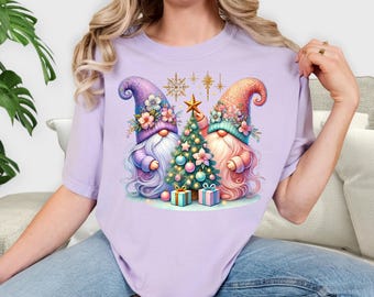 May include: A light purple t-shirt featuring a whimsical Christmas design. The graphic shows two gnomes with floral hats, a decorated Christmas tree, and gift boxes. The word "ORCHID" is printed in black at the bottom of the shirt.