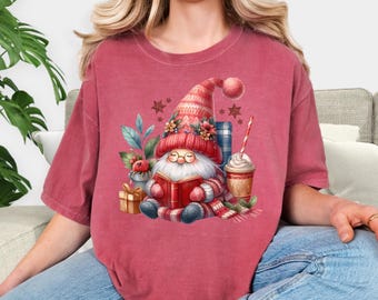 May include: Crimson-colored t-shirt featuring a watercolor illustration of a gnome reading a book. The gnome wears a red hat and scarf, with a coffee cup and gifts nearby. The word "Crimson" is printed at the bottom.