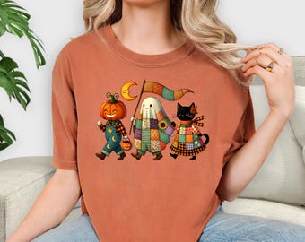 May include: Rust-colored t-shirt featuring a Halloween-themed graphic. The design depicts a pumpkin, a ghost, and a black cat, all dressed in patchwork outfits, walking together. The pumpkin carries a lantern, and the ghost holds a flag with a crescent moon.