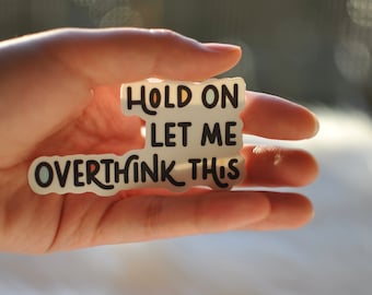 May include: A white sticker with the text "HOLD ON LET ME OVERTHINK THIS" in black and orange lettering. The sticker is held in a hand against a blurred background.