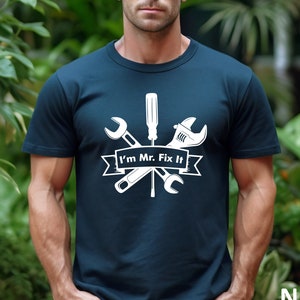 May include: A navy blue t-shirt with a white graphic of tools, including a screwdriver, wrench, and a hammer. The text "I'm Mr. Fix It" is printed on a white banner.