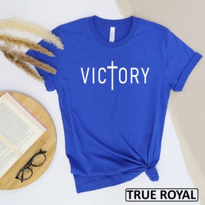 May include: A royal blue t-shirt with the word "VICTORY" printed in white, featuring a cross incorporated into the letter "T". The shirt is made of a soft material and has rolled-up sleeves. The text "TRUE ROYAL" is in a black box at the bottom.