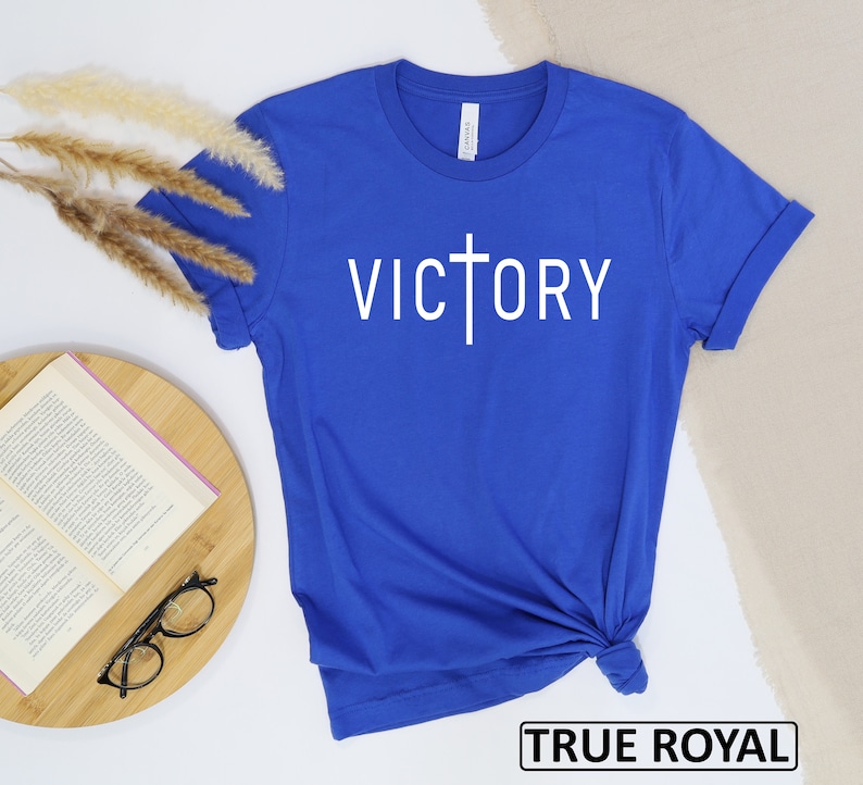May include: A royal blue t-shirt with the word "VICTORY" printed in white, featuring a cross incorporated into the letter "T". The shirt is made of a soft material and has rolled-up sleeves. The text "TRUE ROYAL" is in a black box at the bottom.
