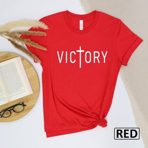 May include: A red t-shirt with the word "VICTORY" printed in white, with a cross incorporated into the letter "T". The shirt is made of a soft material and is displayed on a white surface with a book and glasses.