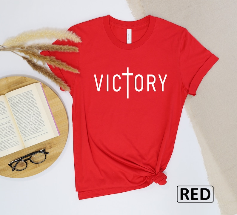 May include: A red t-shirt with the word "VICTORY" printed in white, with a cross incorporated into the letter "T". The shirt is made of a soft material and is displayed on a white surface with a book and glasses.