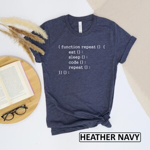 May include: Heather navy t-shirt with white text that reads: (function repeat () { eat (); sleep (); code (); repeat (); })();. The text "HEATHER NAVY" is in a white rectangle at the bottom of the shirt.