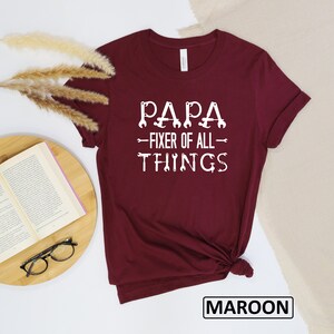 May include: Maroon t-shirt with white text that reads "Papa - Fixer of All - Things" with wrench symbols around the text.