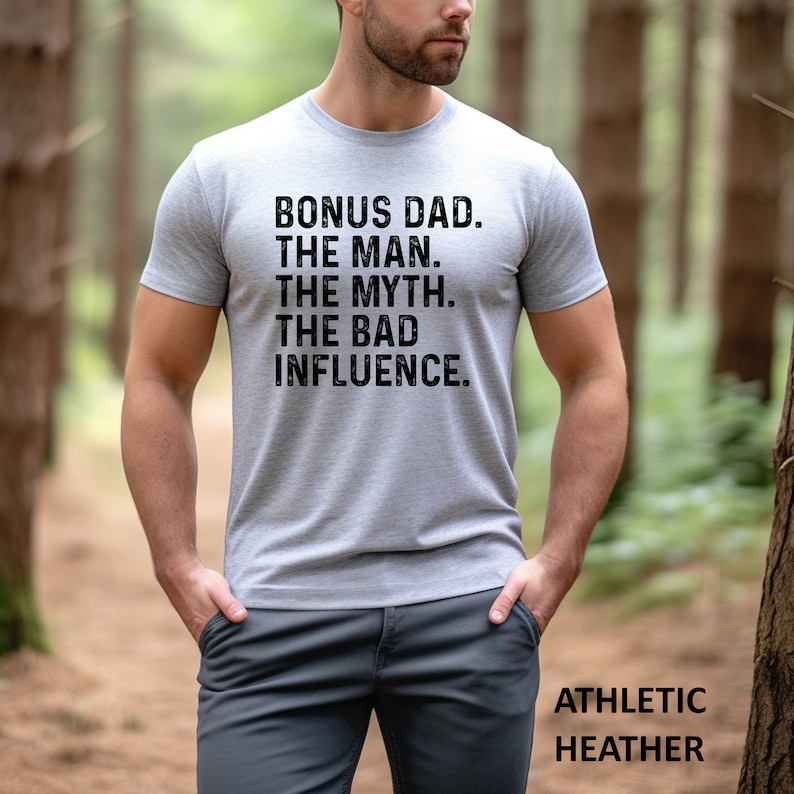 May include: A gray t-shirt with the text "BONUS DAD. THE MAN. THE MYTH. THE BAD INFLUENCE." printed in black. The shirt is shown on a model in a forest setting. The text "ATHLETIC HEATHER" is printed in black at the bottom of the image.