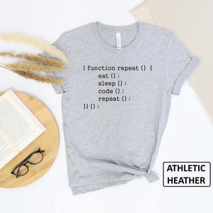 May include: Athletic heather gray t-shirt with the text "(function repeat() { eat(); sleep(); code(); repeat(); })();". The shirt is a crew neck and has a tag that says "ATHLETIC HEATHER".