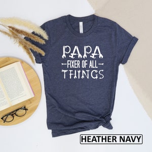 May include: A heather navy blue t-shirt with white text that reads "Papa - Fixer of All Things" with wrench illustrations.