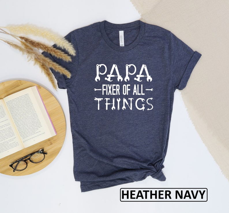 May include: A heather navy blue t-shirt with white text that reads "Papa - Fixer of All Things" with wrench illustrations.
