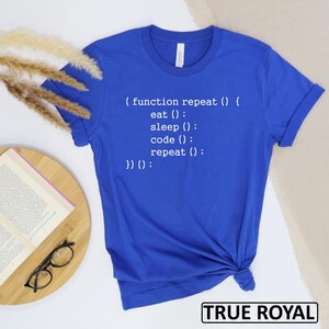 May include: Royal blue t-shirt with white text that reads "(function repeat () { eat (); sleep (); code (); repeat (); })() :". The shirt is folded at the bottom. The words "TRUE ROYAL" are in a black box at the bottom.