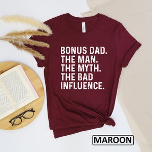 May include: Maroon t-shirt with white text that reads "Bonus Dad. The Man. The Myth. The Bad Influence."