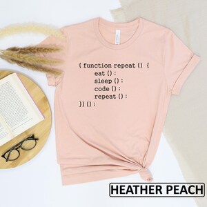 May include: A heather peach-colored t-shirt with black text that reads "(function repeat() { eat(); sleep(); code(); repeat(); })();". The shirt is folded at the bottom. The words "HEATHER PEACH" are in a black rectangle at the bottom.