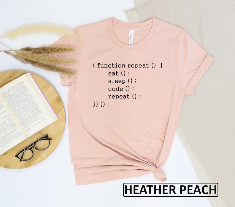 May include: A heather peach-colored t-shirt with black text that reads "(function repeat() { eat(); sleep(); code(); repeat(); })();". The shirt is folded at the bottom. The words "HEATHER PEACH" are in a black rectangle at the bottom.