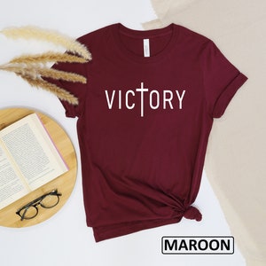 May include: Maroon t-shirt with the word "VICTORY" in white, featuring a cross design. The shirt is folded at the bottom. The word "MAROON" is in a black box at the bottom of the image.