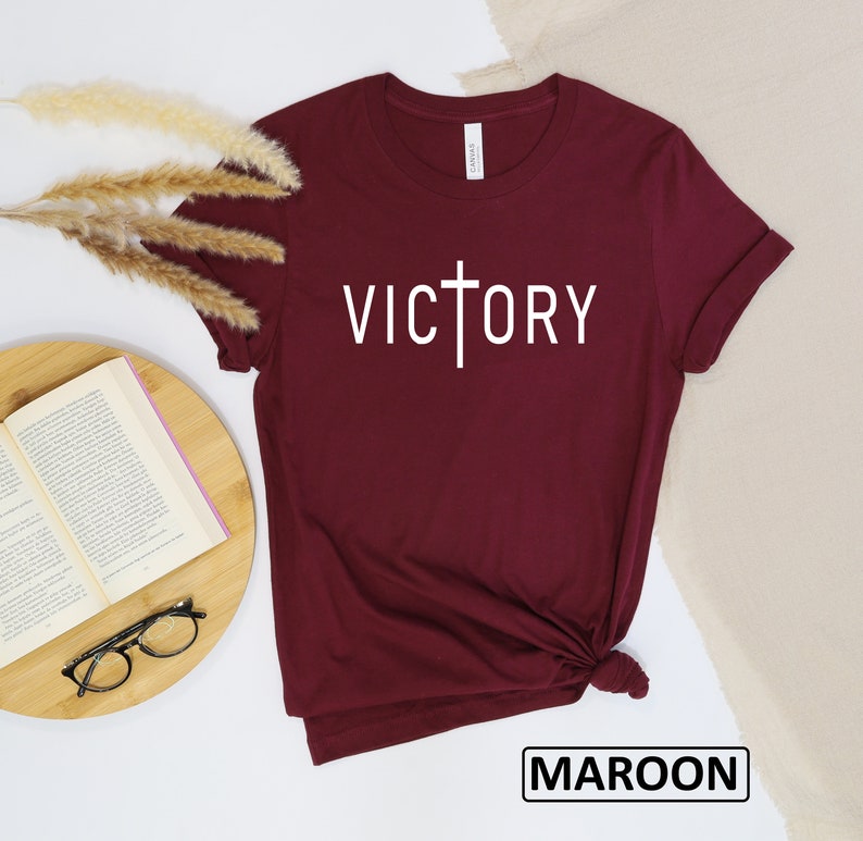 May include: Maroon t-shirt with the word "VICTORY" in white, featuring a cross design. The shirt is folded at the bottom. The word "MAROON" is in a black box at the bottom of the image.