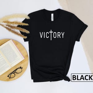 May include: Black t-shirt with the word "VICTORY" in white, featuring a cross design. The shirt is displayed on a wooden surface with a book, glasses, and decorative dried plants. The word "BLACK" is in a white box.