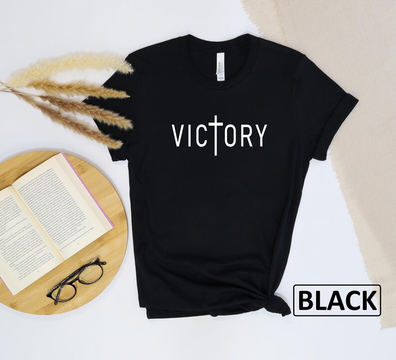 May include: Black t-shirt with the word "VICTORY" in white, featuring a cross design. The shirt is displayed on a wooden surface with a book, glasses, and decorative dried plants. The word "BLACK" is in a white box.