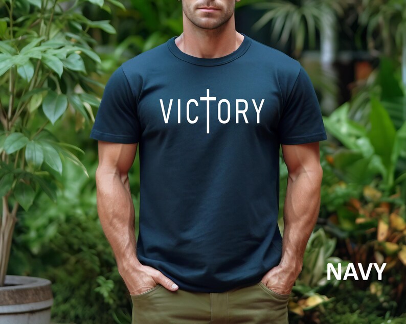 May include: Navy blue t-shirt with the word "VICTORY" in white, featuring a cross design. The shirt is a crew neck style and is made of a soft material. The word "NAVY" is visible in the bottom right corner.