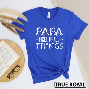 May include: Royal blue t-shirt with white text that reads "Papa - Fixer of All Things" with wrench icons. The text is centered on the shirt.