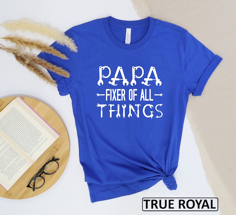 May include: Royal blue t-shirt with white text that reads "Papa - Fixer of All Things" with wrench icons. The text is centered on the shirt.
