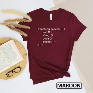 May include: Maroon t-shirt with white text that reads "(function repeat () { eat (); sleep (); code (); repeat (); })() :". The shirt is a solid color and has a crew neck. The word "MAROON" is printed in a white box at the bottom.