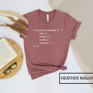 May include: Heather mauve t-shirt with the text "(function repeat() { eat(); sleep(); code(); repeat(); })();" in white. The shirt is folded and has a rolled sleeve. The words "HEATHER MAUVE" are in a black box.
