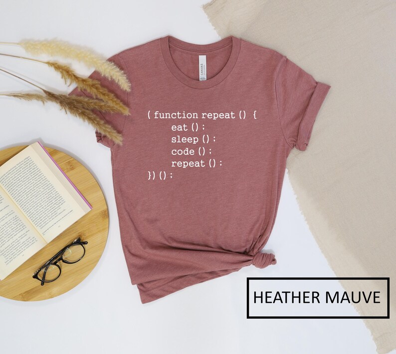 May include: Heather mauve t-shirt with the text "(function repeat() { eat(); sleep(); code(); repeat(); })();" in white. The shirt is folded and has a rolled sleeve. The words "HEATHER MAUVE" are in a black box.