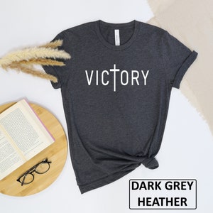 May include: Dark grey heather t-shirt with the word "VICTORY" in white, featuring a cross design. The shirt is folded, with a book, glasses, and decorative plant in the background. The text "DARK GREY HEATHER" is in a black rectangle.