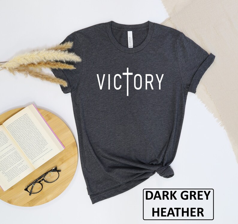 May include: Dark grey heather t-shirt with the word "VICTORY" in white, featuring a cross design. The shirt is folded, with a book, glasses, and decorative plant in the background. The text "DARK GREY HEATHER" is in a black rectangle.