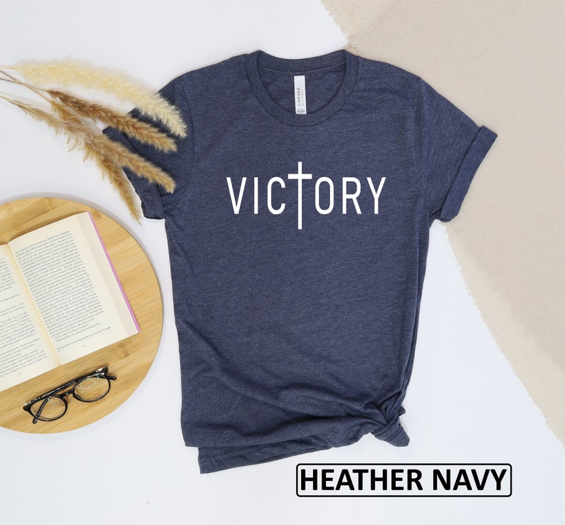 May include: Heather navy t-shirt with the word "VICTORY" in white, featuring a cross design. The shirt is made of a soft material and has rolled-up sleeves. The text "HEATHER NAVY" is printed at the bottom.