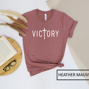 May include: Heather mauve t-shirt with the word "VICTORY" in white, featuring a cross design. The shirt is folded at the bottom and has rolled-up sleeves. The text "HEATHER MAUVE" is in a black box at the bottom.
