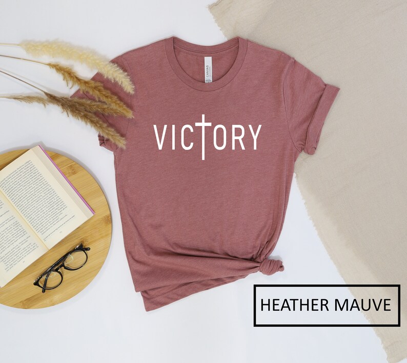 May include: Heather mauve t-shirt with the word "VICTORY" in white, featuring a cross design. The shirt is folded at the bottom and has rolled-up sleeves. The text "HEATHER MAUVE" is in a black box at the bottom.