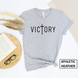 May include: Athletic heather gray t-shirt with the word "VICTORY" printed in black, with a cross replacing the "T". The shirt is displayed on a white surface with a book, glasses, and decorative plants.