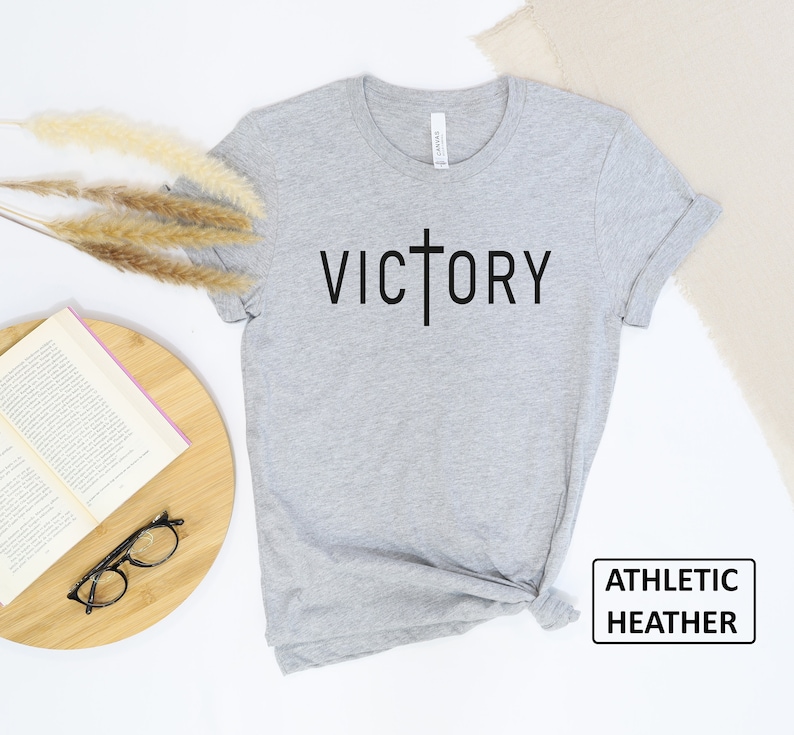 May include: Athletic heather gray t-shirt with the word "VICTORY" printed in black, with a cross replacing the "T". The shirt is displayed on a white surface with a book, glasses, and decorative plants.