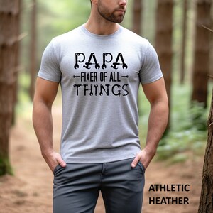 May include: A gray t-shirt with the text "PAPA - FIXER OF ALL - THINGS" in black. The text is surrounded by wrench and screwdriver images. The shirt is being worn by a person in a forest setting.