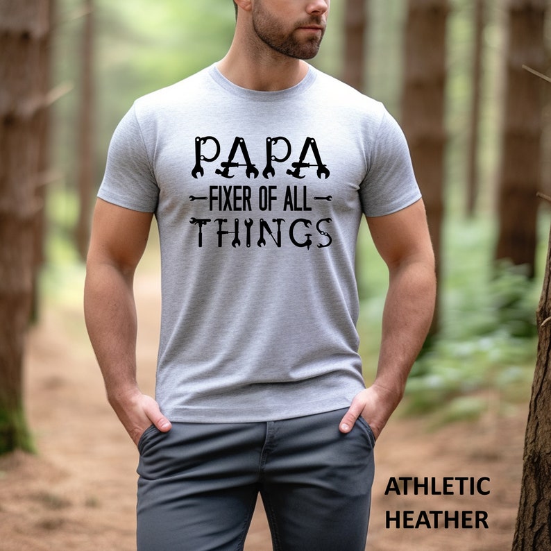 May include: A gray t-shirt with the text "PAPA - FIXER OF ALL - THINGS" in black. The text is surrounded by wrench and screwdriver images. The shirt is being worn by a person in a forest setting.