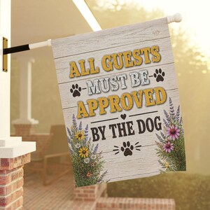May include: A rectangular wooden-style house flag with the text "ALL GUESTS MUST BE APPROVED BY THE DOG". The flag has a light wood grain background with yellow and white text, paw prints, and floral accents.