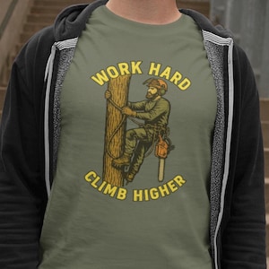 May include: Olive green t-shirt featuring a graphic of a lumberjack climbing a tree with a chainsaw. The text "WORK HARD" is arched above the illustration, and "CLIMB HIGHER" is arched below. The shirt is worn by a person.